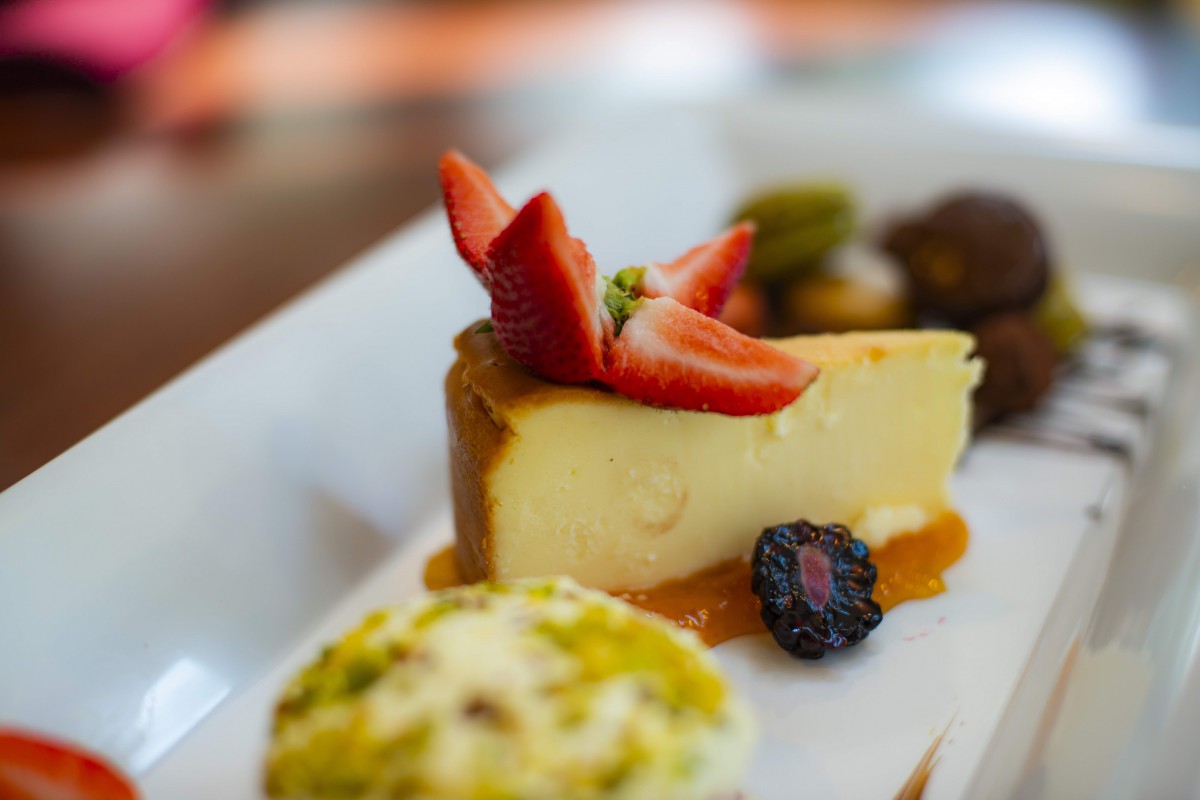 A delicious slice of cheesecake topped with fresh strawberries, accompanied by a pistachio dessert and a blackberry on the side.