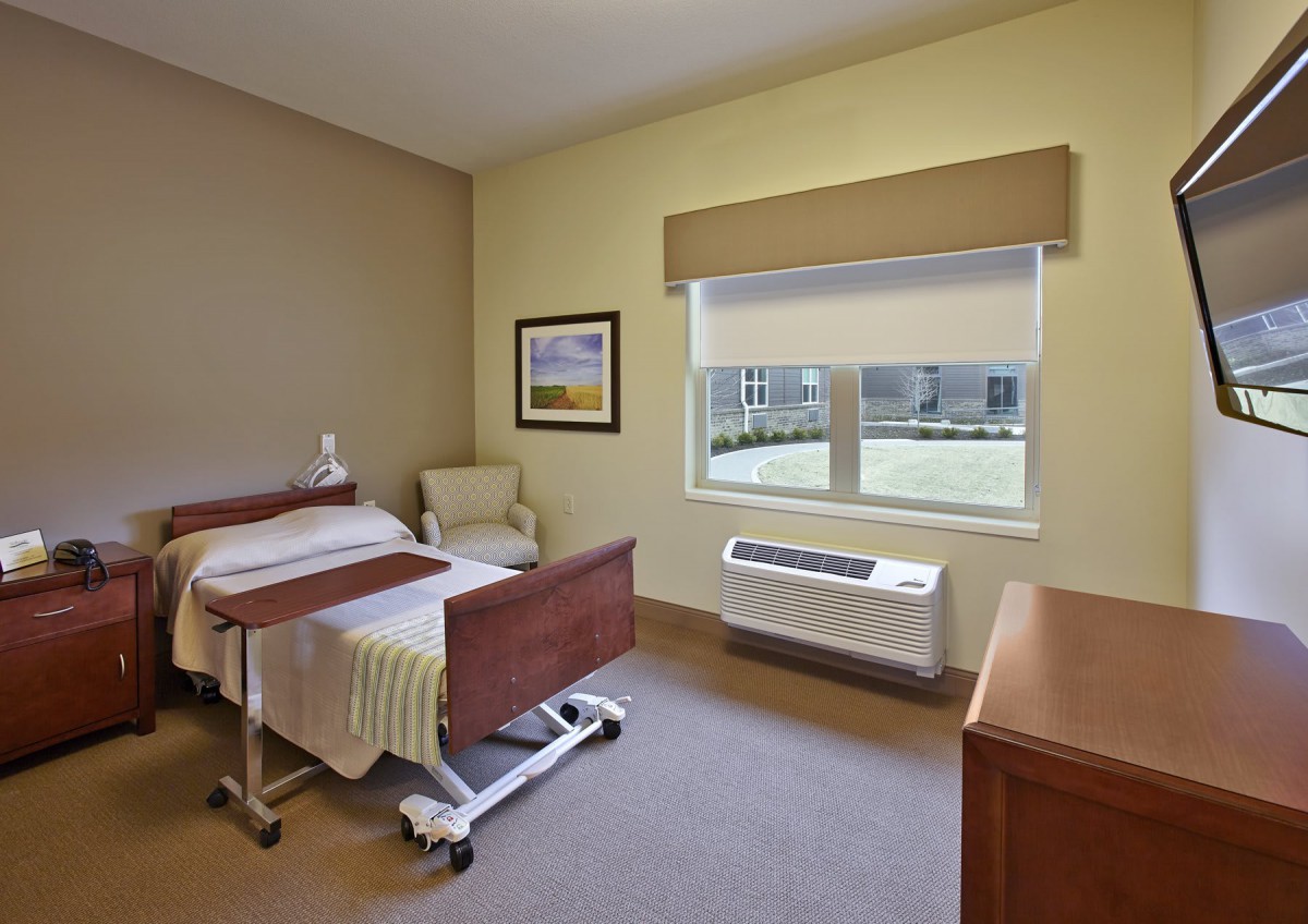 A simple and clean hospital room with a bed, bedside table, chair, and a window overlooking a garden.