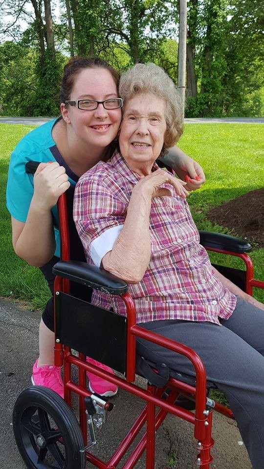 A younger woman hugging an elderly woman in a wheelchair outdoors.