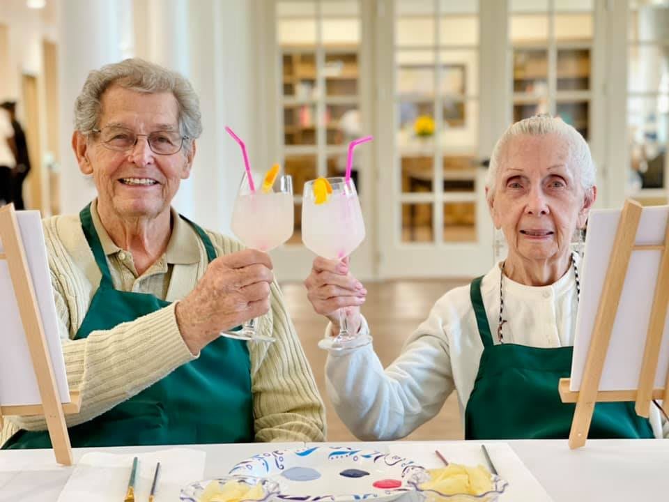 An elderly couple toasting with drinks while smiling indoors.