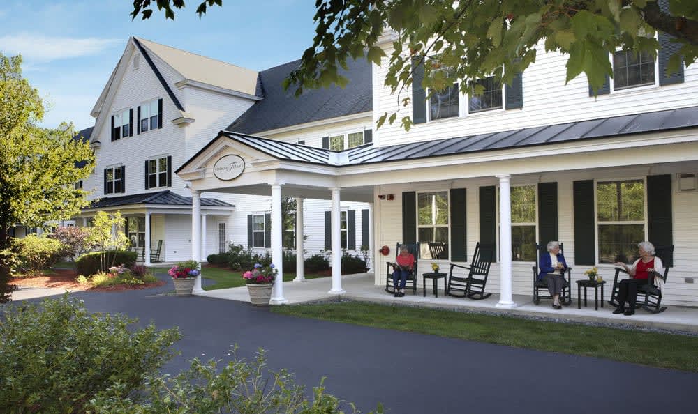 A white building with a porch where people are seated, surrounded by trees.