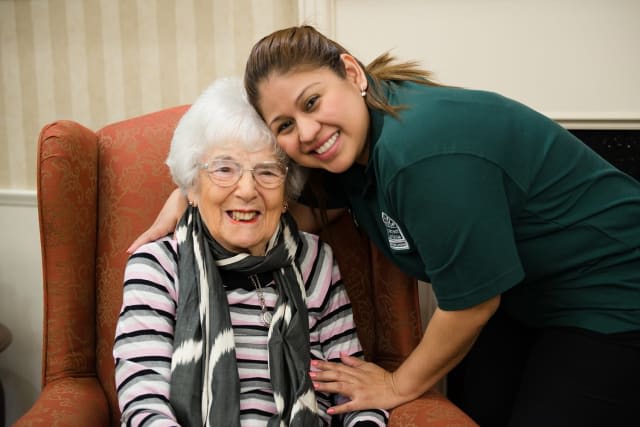 An elderly woman sitting and smiling with a caregiver beside her.