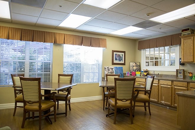 A cozy dining area with tables and chairs, well-lit by large windows and ceiling lights.