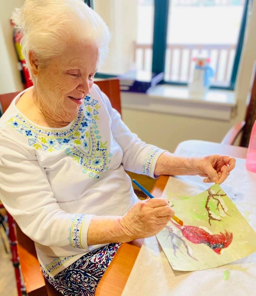An elderly woman enjoys painting indoors, engaging in a creative activity.