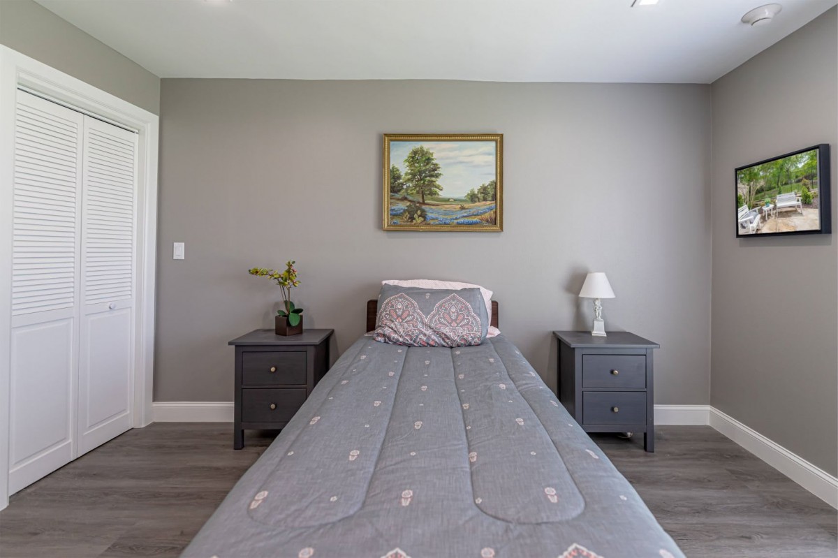 A minimalist bedroom featuring a single bed with gray bedding, flanked by two nightstands and a lamp, against soft gray walls adorned with a landscape painting.