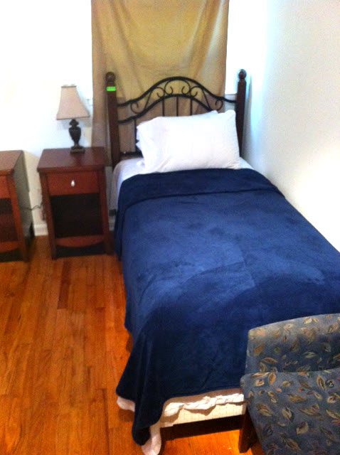 A cozy bedroom with a single bed featuring a blue blanket, accompanied by two nightstands and a lamp on a wooden floor.