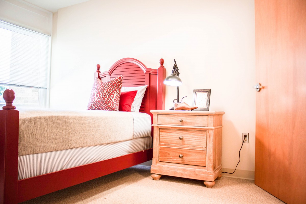 A cozy bedroom featuring a red bed with decorative pillows and a wooden nightstand with a lamp.