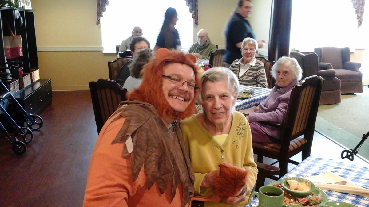 A person in a costume posing with an elderly woman at a group gathering indoors.