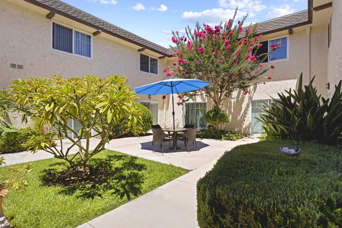 A peaceful courtyard in an apartment complex with outdoor seating and a blue umbrella surrounded by lush greenery.