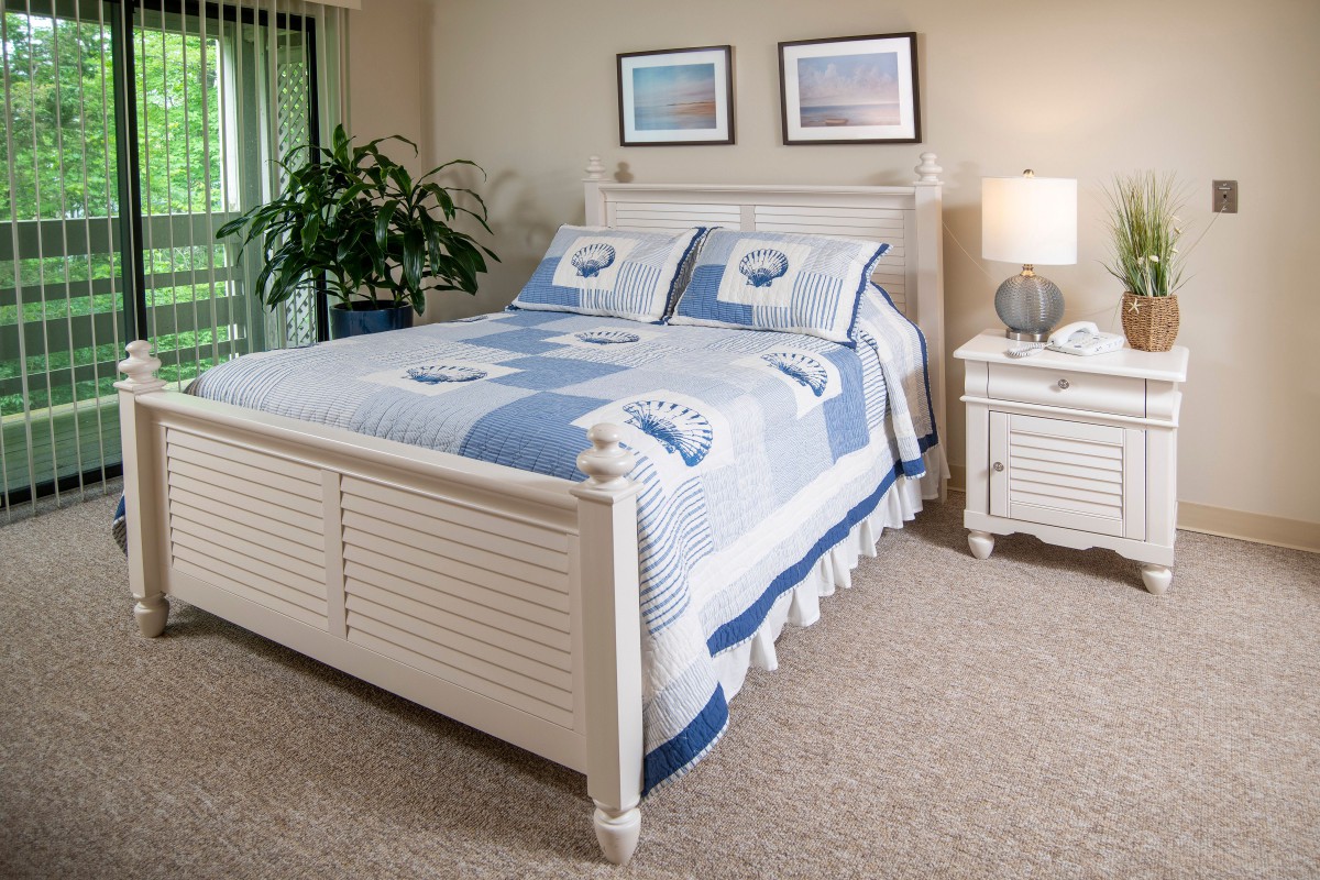 A cozy bedroom with a blue and white bedspread, nightstand, lamp, and framed artwork on the wall.