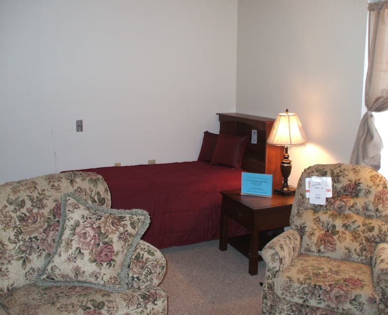 A cozy bedroom setup featuring a bed with burgundy bedding, a wooden nightstand with a lamp, and floral-patterned armchairs.