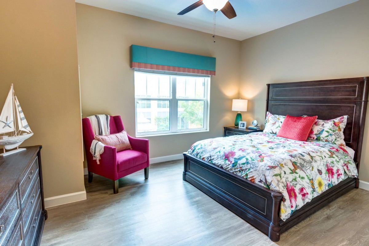 A cozy bedroom with a colorful bedspread, a pink armchair, and natural light from a window.