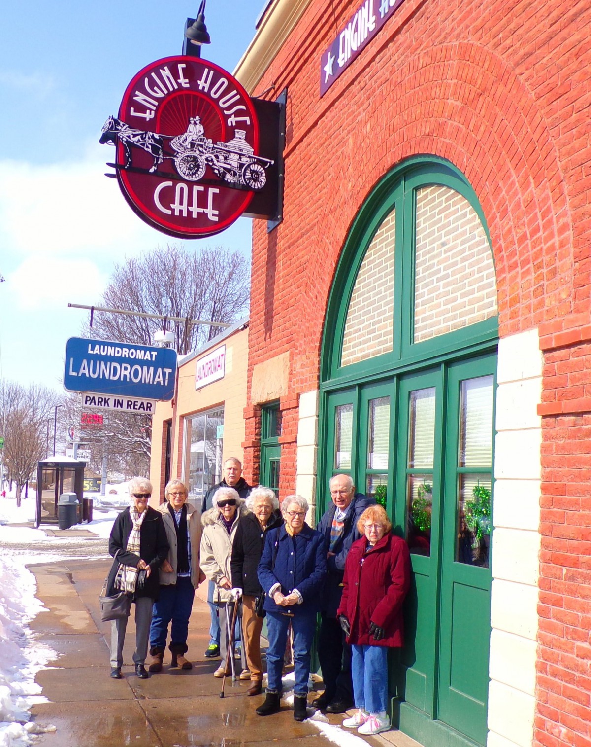 A group of people standing outside a caf with a distinctive sign, on a snowy day.