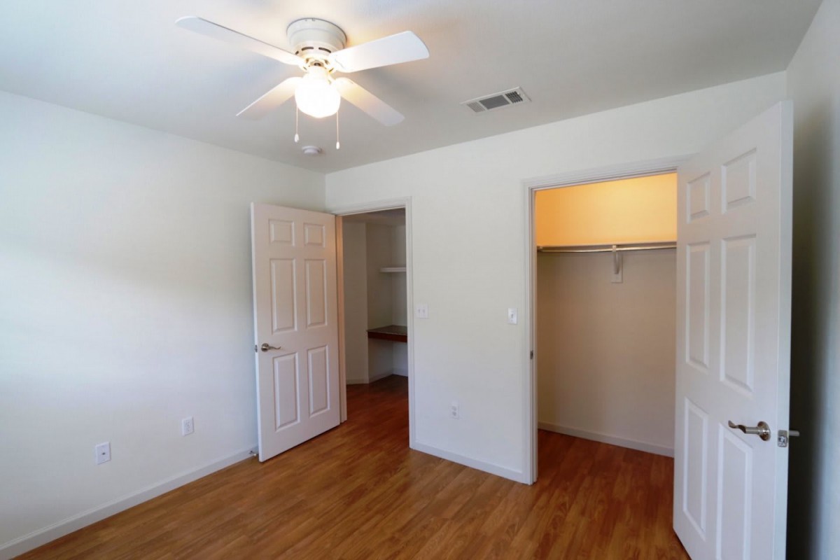 A bright, empty room with wooden floors and a ceiling fan, featuring an open closet.