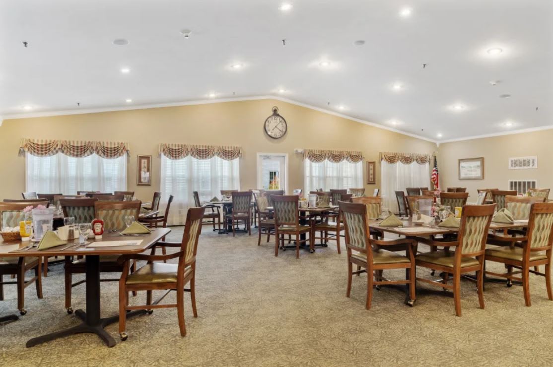 A spacious and well-lit dining area with multiple tables and chairs, featuring neutral decor and a central clock on the wall.