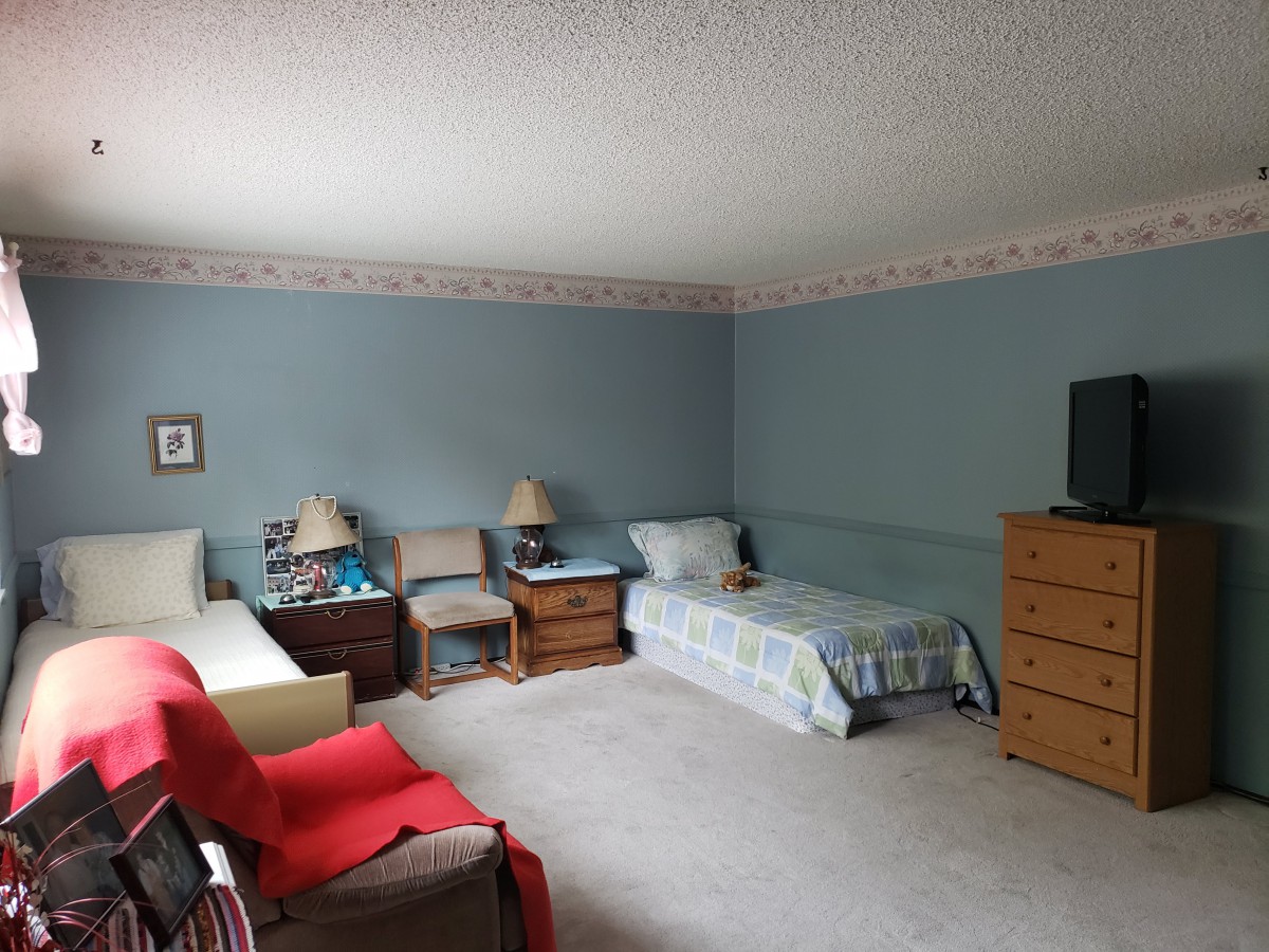 A cozy bedroom with blue walls, featuring a single bed, a dresser, armchairs, and a television.