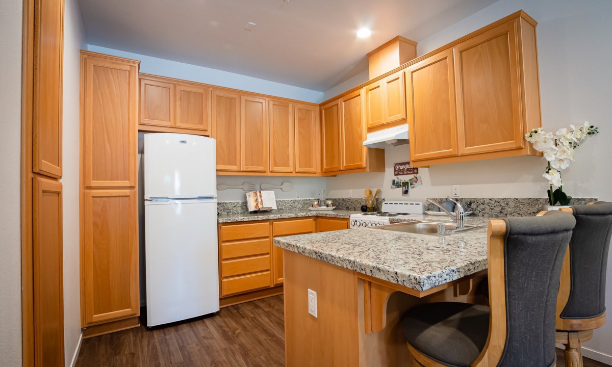 A modern kitchen with wooden cabinets and a granite countertop, featuring a white refrigerator and stovetop.