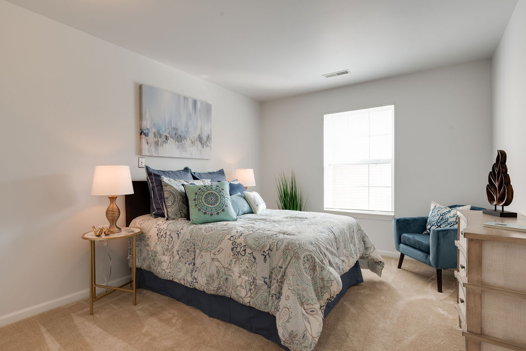 A cozy and elegantly decorated bedroom featuring a neatly made bed with patterned bedding, side tables with lamps, a piece of artwork above the bed, and a window letting in natural light.