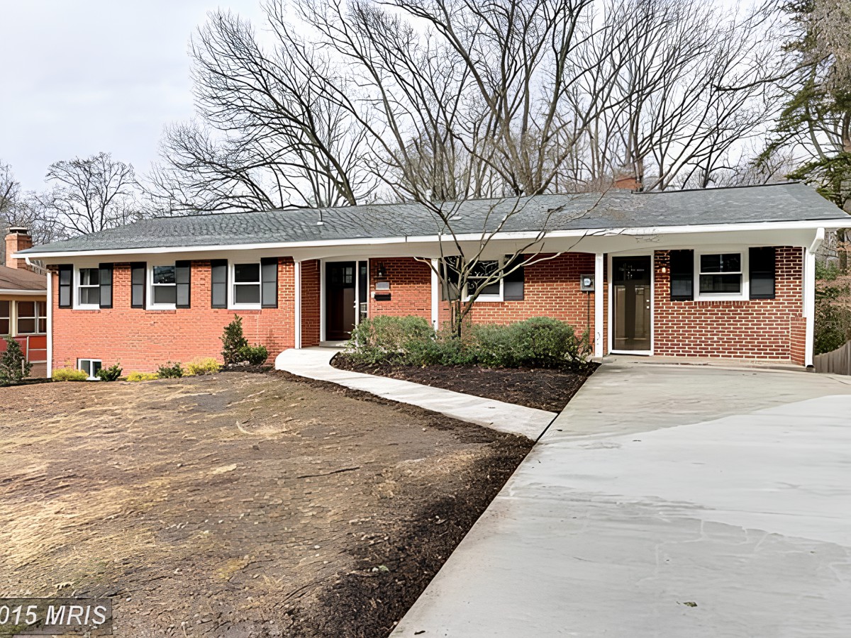 A suburban brick house with a concrete driveway and surrounding trees.