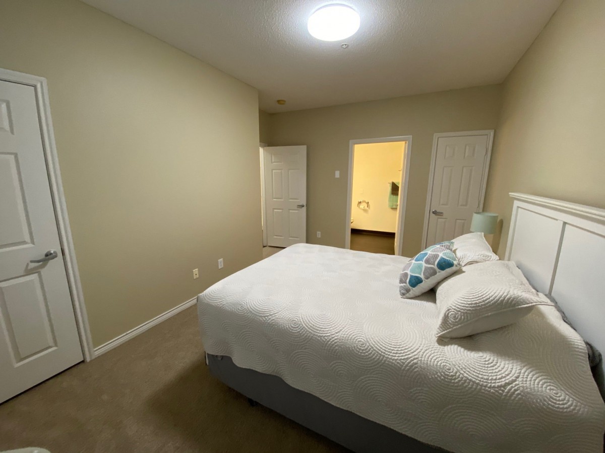 A minimalist bedroom with a neatly made bed, carpet flooring, and two doors leading to other rooms.