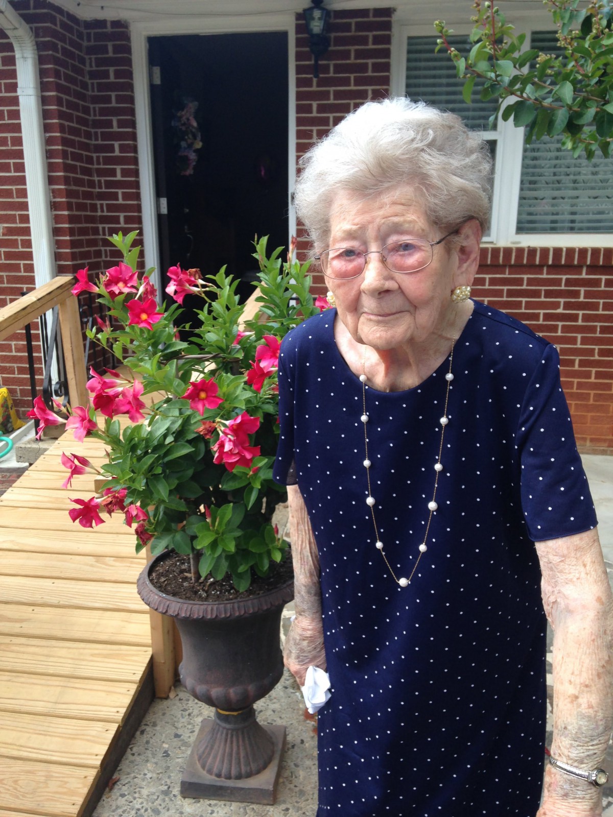 An elderly woman standing next to a pot of vibrant flowers in an outdoor garden setting.