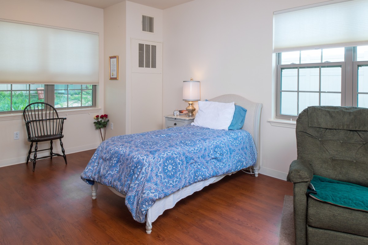 A cozy bedroom with a single bed adorned with a blue bedspread, complemented by wooden flooring and a lamp on the nightstand.