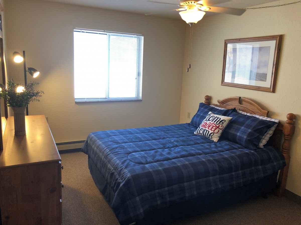 A cozy bedroom featuring a bed with blue plaid bedding, a wooden dresser, and a ceiling fan.
