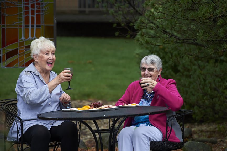 Two elderly women enjoying drinks and socializing outdoors at a table in a garden.
