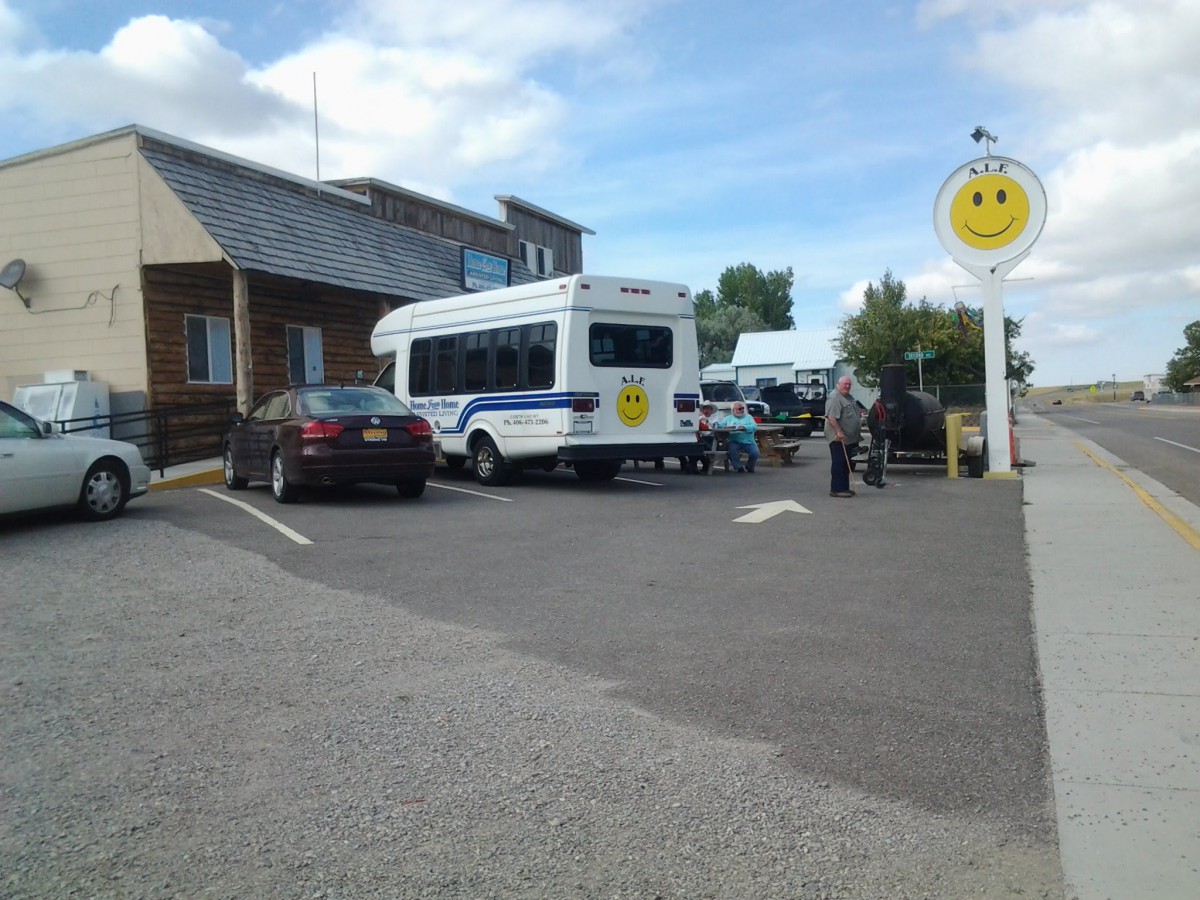 A small bus and several cars are parked in a parking lot near a building, with a smiley face sign visible against the blue sky.