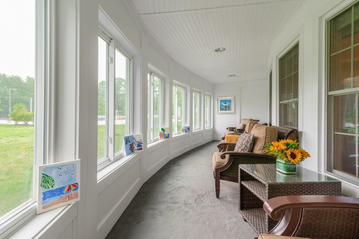 A bright sunroom with large windows, featuring comfortable chairs and a table with flowers.