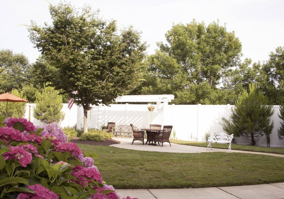 A serene backyard garden with outdoor furniture set on a patio, surrounded by lush trees and blooming flowers.