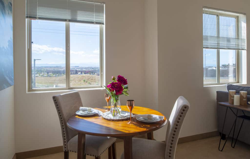 A cozy dining area with a round table set for two, featuring a vase of flowers next to a sunny window.
