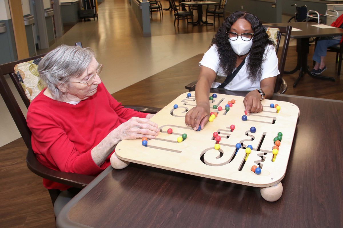 A caregiver and an elderly woman engage with an activity board in a nursing home setting.