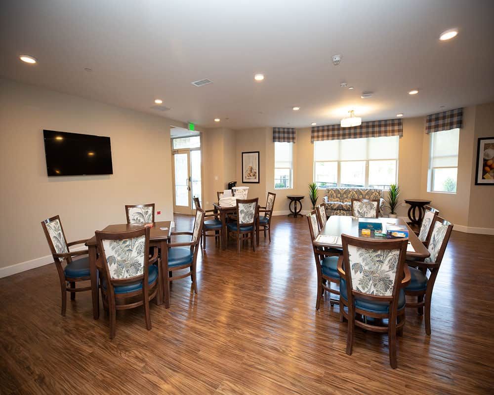 A spacious dining room with multiple tables and chairs, featuring a television and large windows.