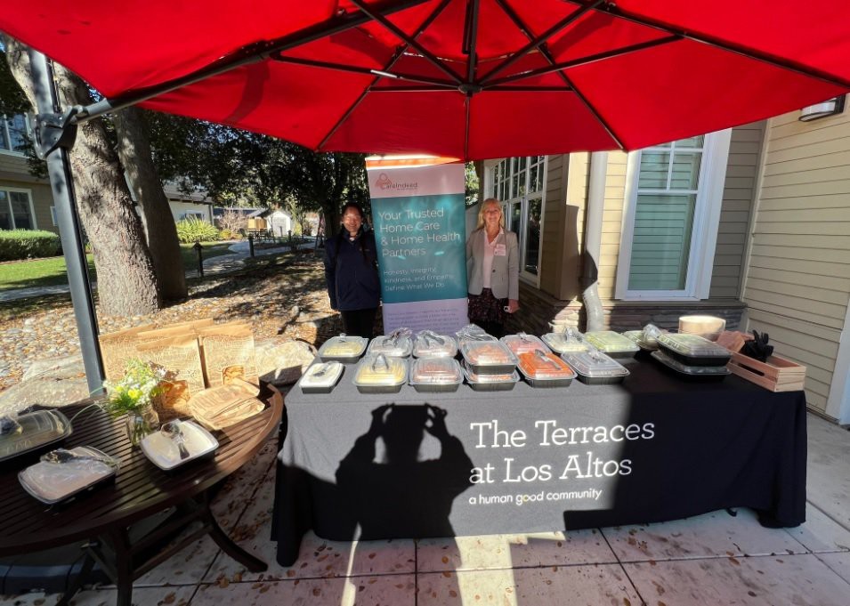 An outdoor event showcasing a variety of food dishes on a table, covered by a large red umbrella. Two people stand behind the display at The Terraces at Los Altos.