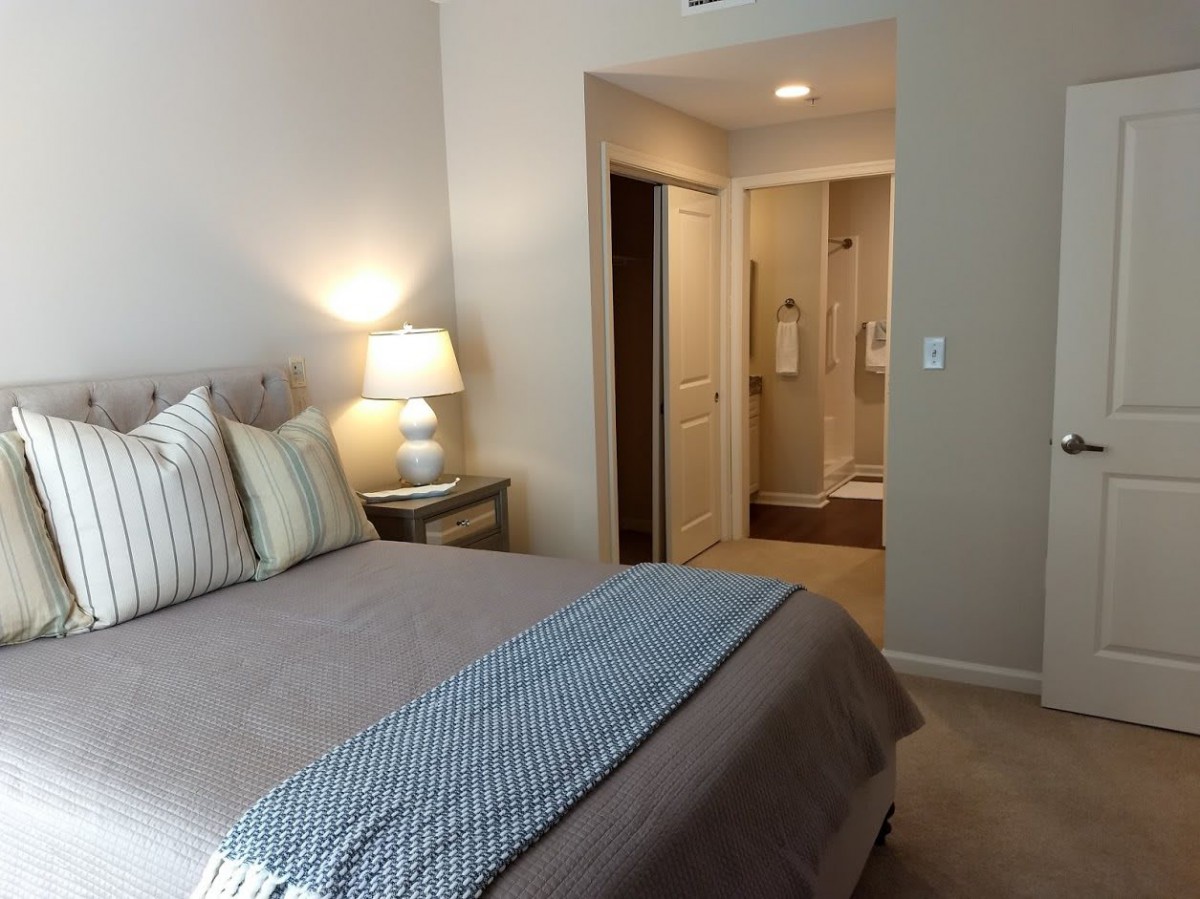 A cozy bedroom with neutral tones, featuring a neatly made bed, side lamp, and an open doorway leading to the bathroom.