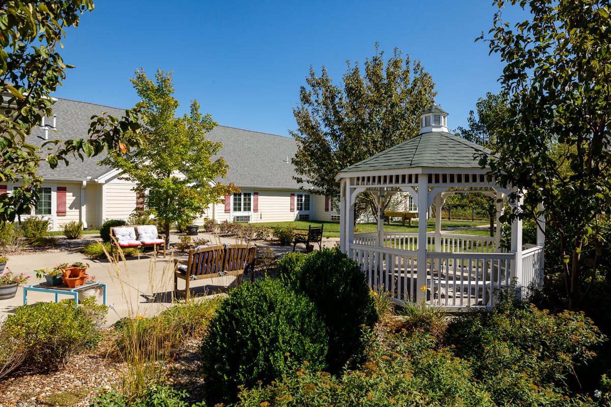 A sunny day view of a gazebo in a well-maintained garden with trees and a house in the background.