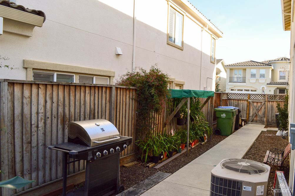 A backyard with a grill, a small garden area, and trash bins along a wooden fence.