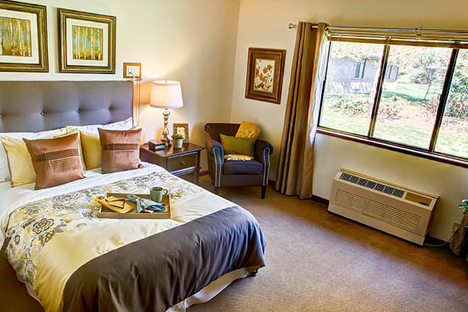 A cozy bedroom with a neatly made bed, side table, and a comfortable chair by the window.