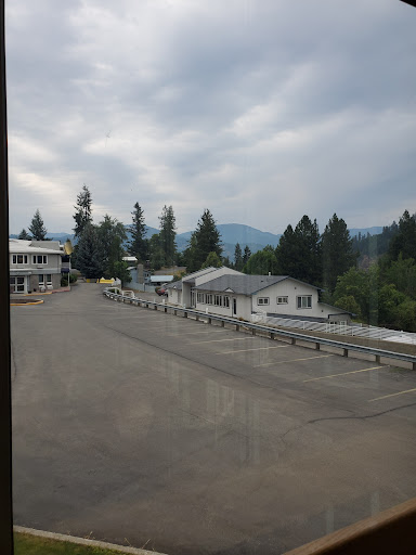 A quiet rural scene featuring buildings along a road, framed by trees and mountains under a cloudy sky.