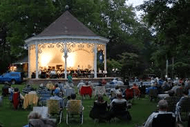 An outdoor evening concert in a park with an audience seated near a gazebo.