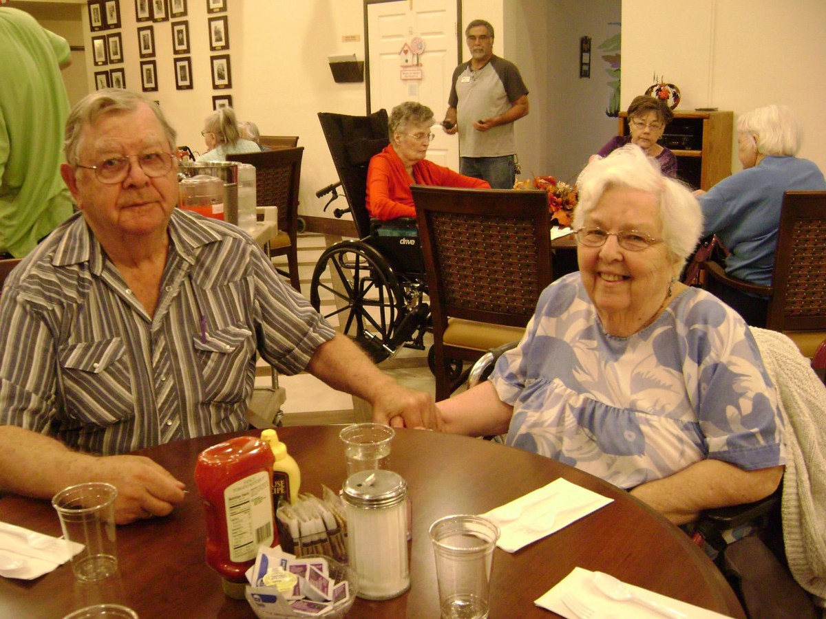 An elderly couple is sitting together at a dining table, holding hands in a cozy restaurant setting.