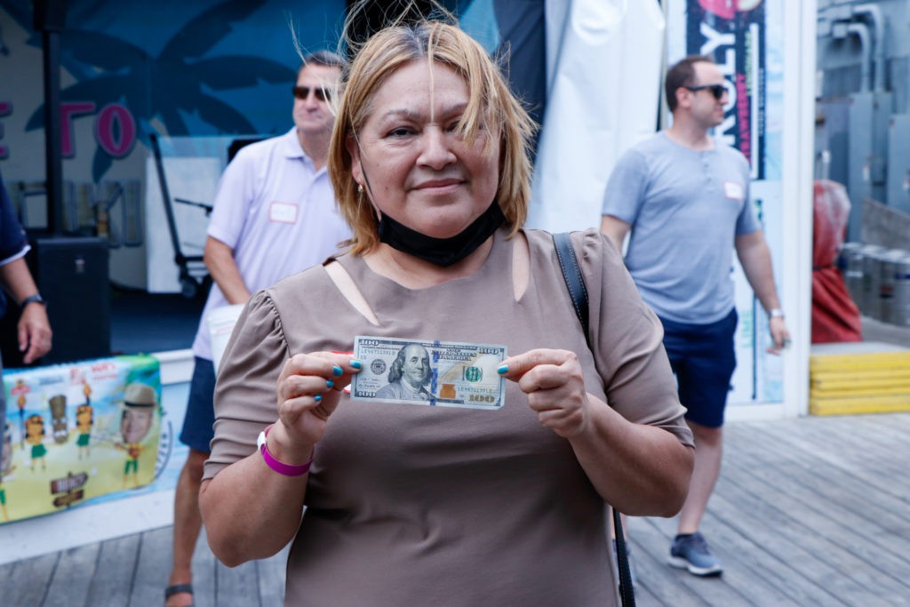 A woman standing outdoors holding a hundred-dollar bill, with people in the background.