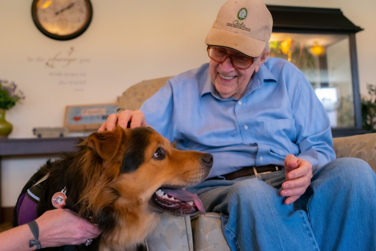 An elderly man sits indoors, smiling warmly as he pets a large dog.