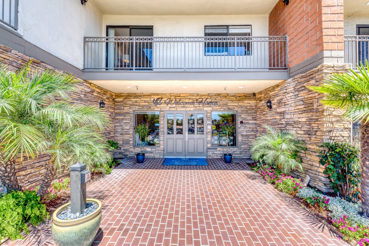 A modern building entrance with brick flooring and decorative stone walls, flanked by lush plants.
