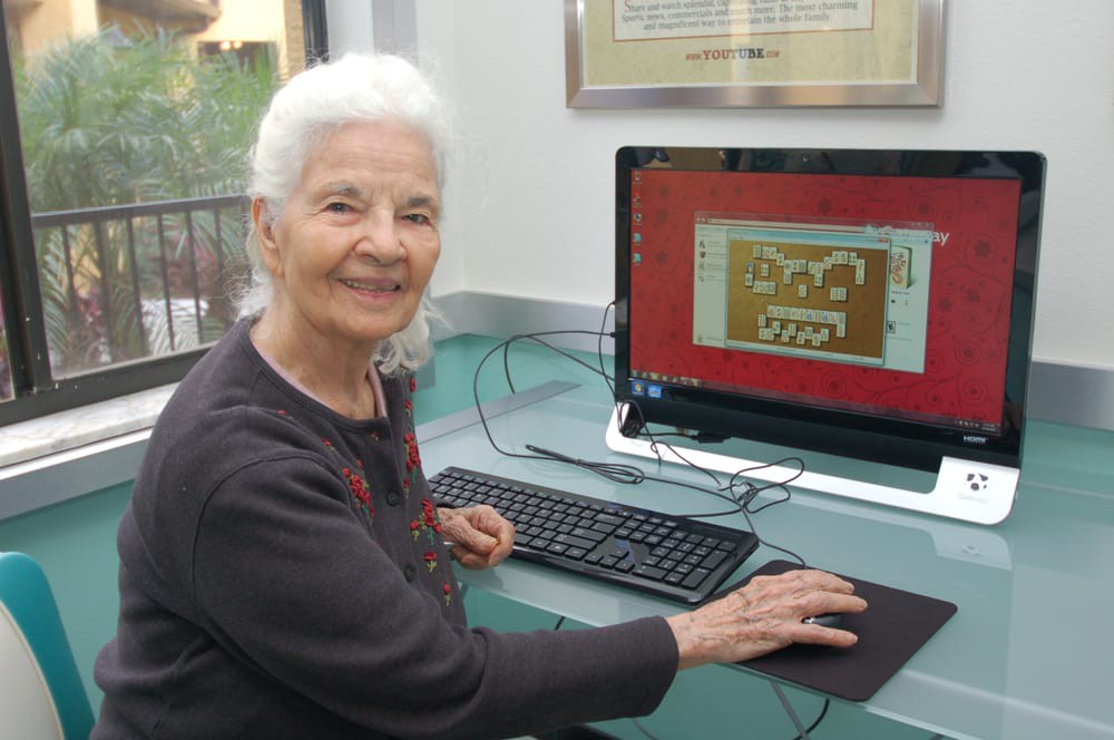 An elderly woman smiling while using a computer in an office setting.