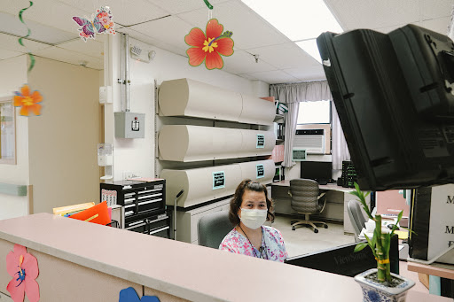 A healthcare worker at the reception desk in a hospital, wearing a mask and surrounded by office equipment.