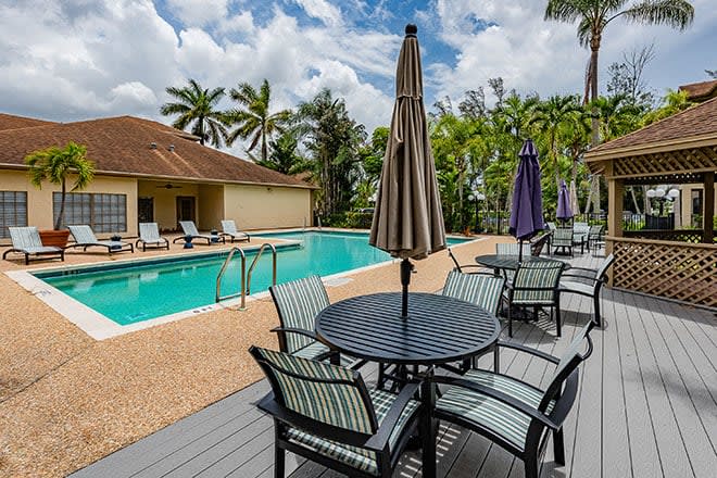 A tranquil outdoor swimming pool area with patio seating and umbrellas, surrounded by lush palm trees.