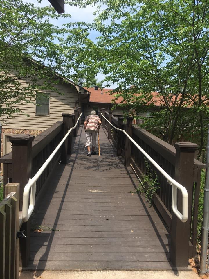 A person walks on a wooden bridge leading to a rustic building surrounded by trees.