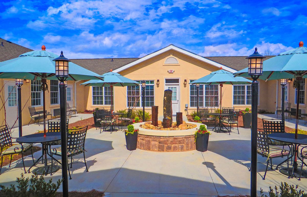 A sunny outdoor patio with a central fountain, surrounded by chairs and umbrellas, against a backdrop of a building and blue sky.